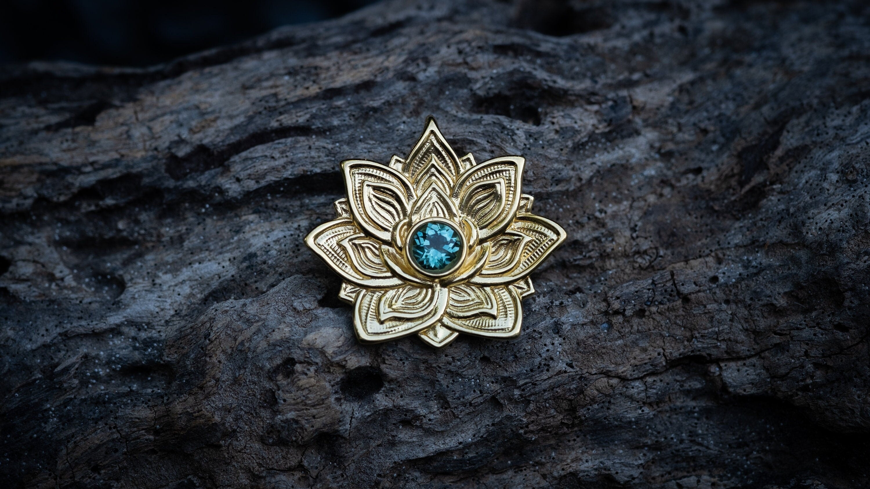 A 14k gold lotus flower pendant with intricately carved petals surrounds a blue gemstone at its center, resting on a piece of driftwood.