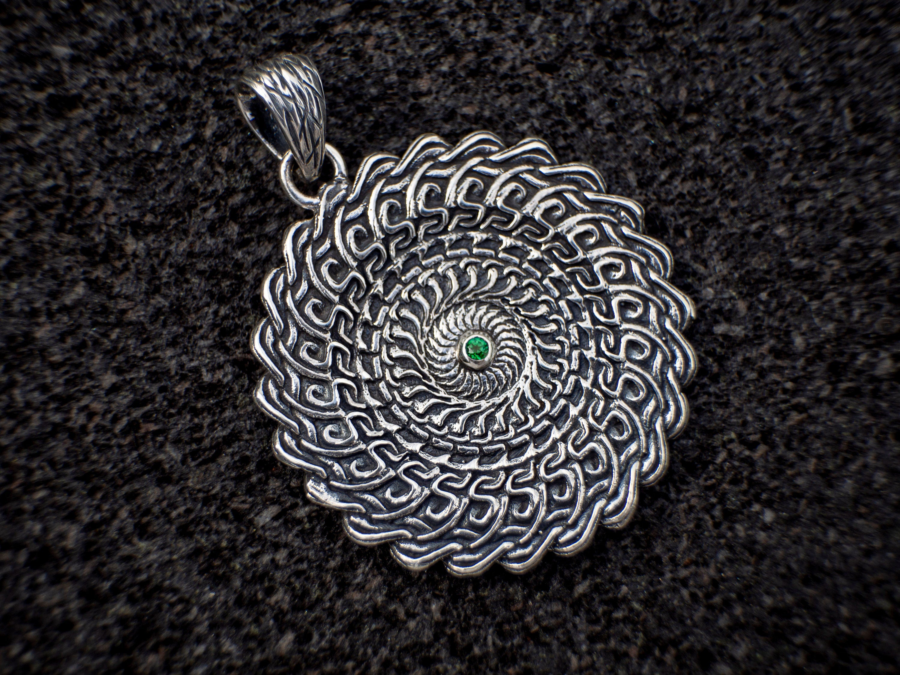 The back of the silver pendant features an intricate mandala design with a green gemstone at the center. The pendant is displayed on a piece of black volcanic rock, highlighting its unique details.